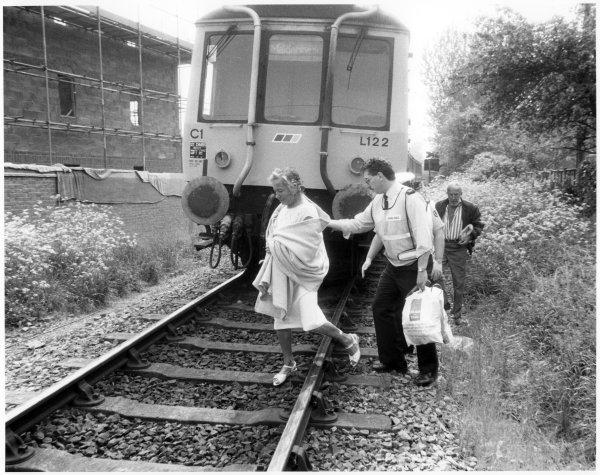 A woman is being helped across a railway line by an ambulance-man in front of a stationary train, with a man behind, Bourne End, Wooburn, circa 1970