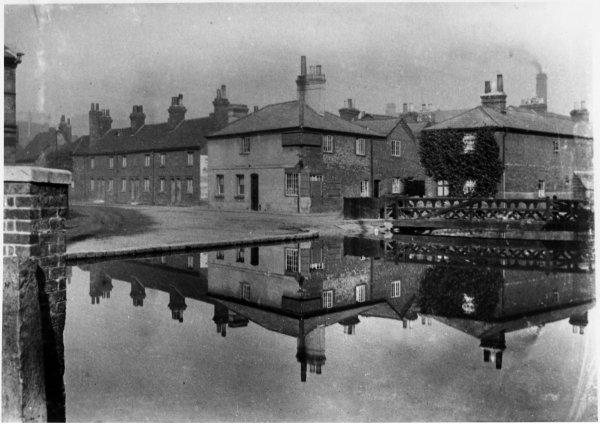 View of the Elephant and Castle pub, the almshouses, and footbridge over the river Wye, all reflected in the river at Newland, High Wycombe. c1895