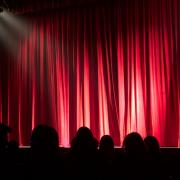 A crowd sitting in front of a theatre red curtain. Credit: Canva