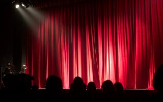 A crowd sitting in front of a theatre red curtain. Credit: Canva